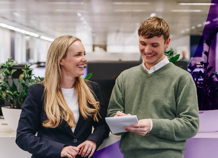 Two colleagues, a man and a woman, smiling and engaging in a conversation while looking at a notepad in a modern office setting.