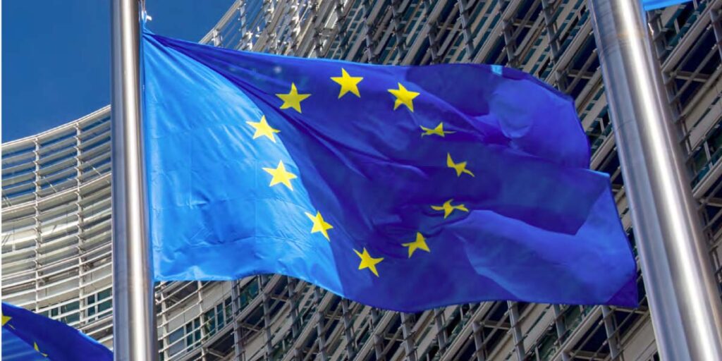 European Union flag with twelve golden stars on a blue background, waving on a flagpole in front of a modern glass-and-steel building