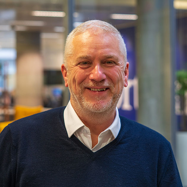 Smiling man with short gray hair wearing a navy sweater in a modern office setting.