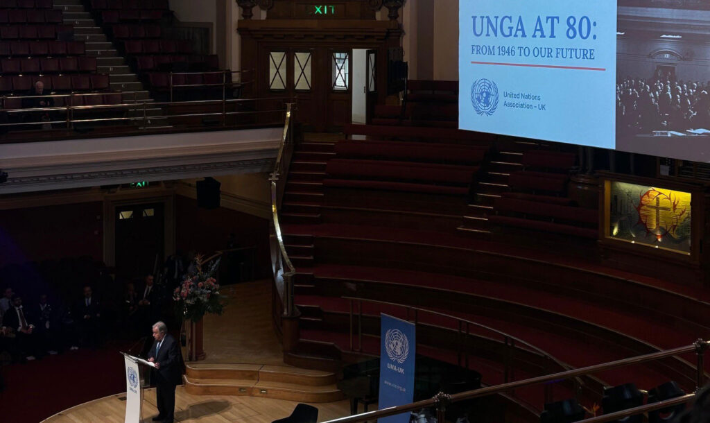 Large, ornate auditorium with tiered red seating and a high domed ceiling. A speaker stands at a podium on a wooden stage. A large screen displays the text “UNGA at 80: From 1946 to Our Future, 17 January 2026,” with United Nations Association–UK branding.