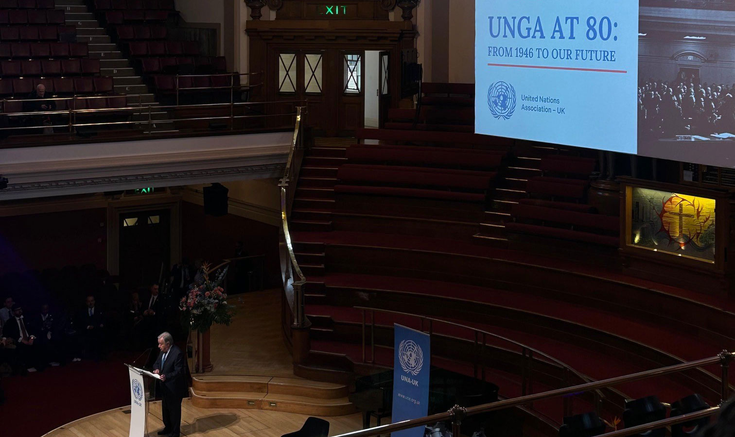 Large, ornate auditorium with tiered red seating and a high domed ceiling. A speaker stands at a podium on a wooden stage. A large screen displays the text “UNGA at 80: From 1946 to Our Future, 17 January 2026,” with United Nations Association–UK branding.