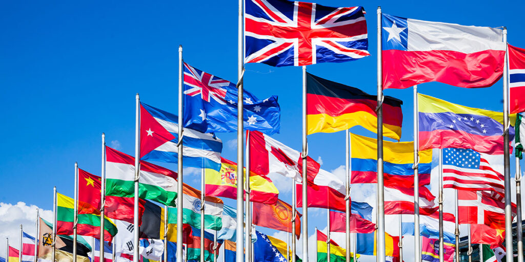 Multiple international flags waving on tall poles against a clear blue sky.