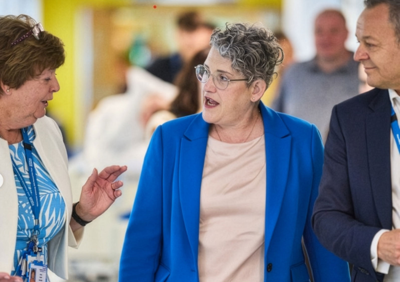 Three adults walk and talk together down a brightly lit hospital corridor. The person in the center wears a blue suit and glasses and appears to be speaking, while the two people beside them listen attentively. Hospital staff and beds are visible in the blurred background, suggesting an active clinical setting.