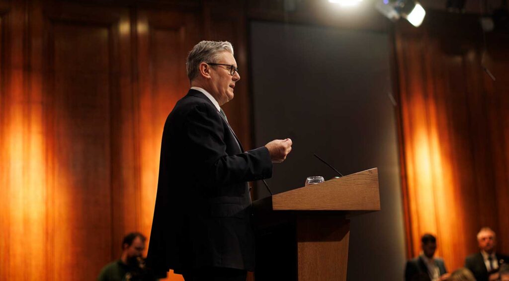 Keir Starmer in a dark suit and glasses speaks at a podium in a warmly lit, wood-paneled room, gesturing with one hand while addressing an audience seated in the background.
