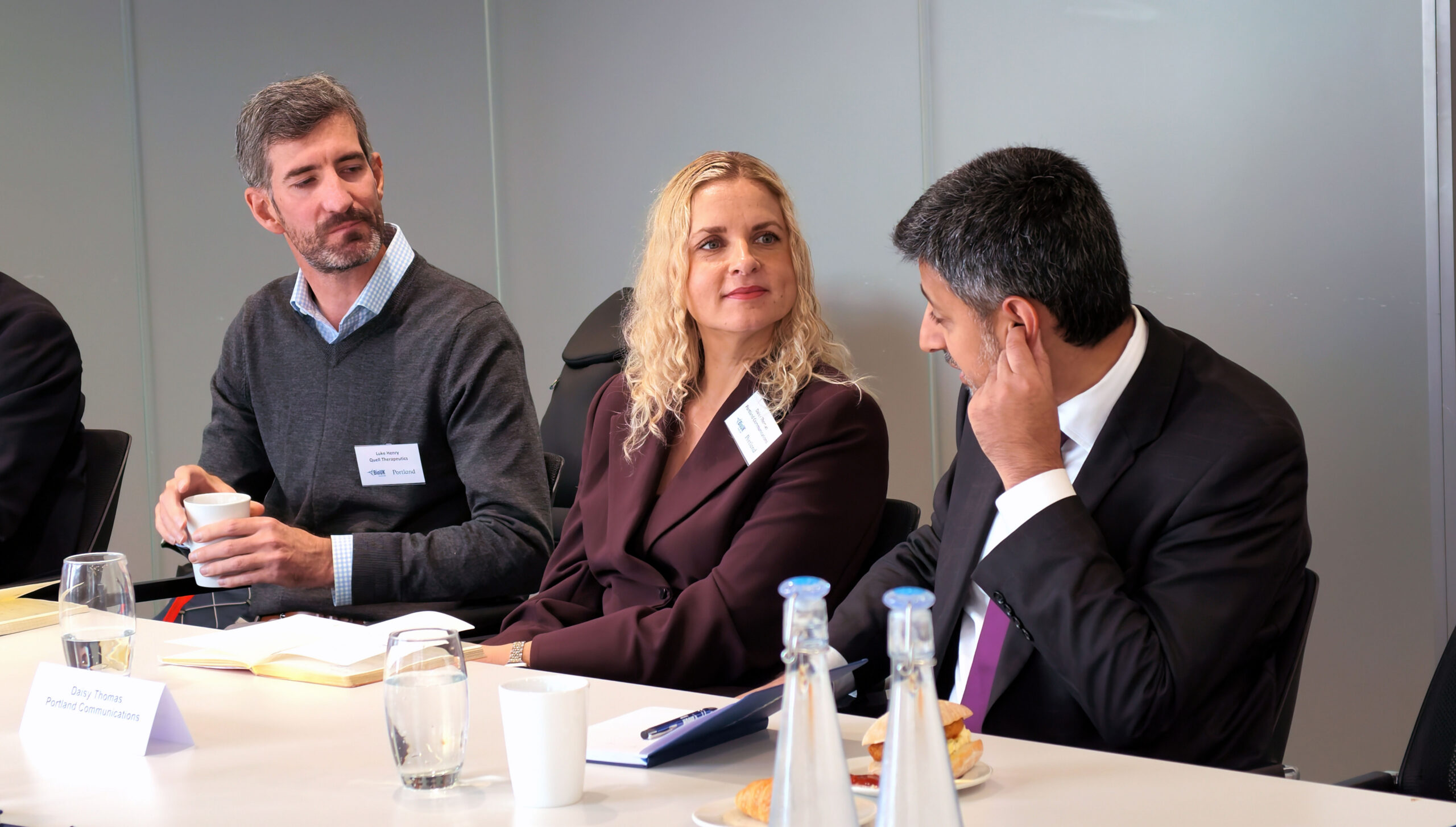 Three people sit together at a networking event, wearing name badges and holding drinks.